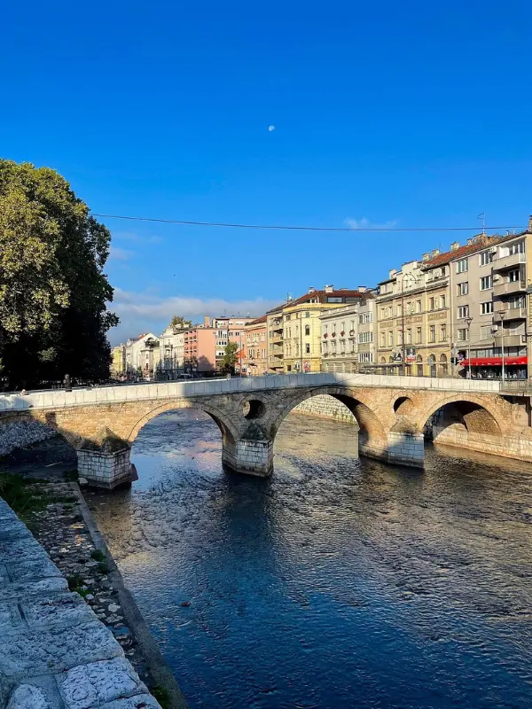 A historic stone arch bridge crossing the Miljacka River with old Sarajevo buildings in the background under a clear blue sky.