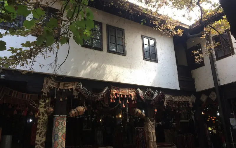 A traditional Ottoman-era inn courtyard with hanging carpets and handicrafts for sale, shaded by trees.