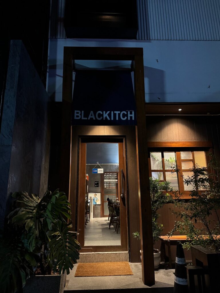 Entrance of Blackitch Artisan Kitchen at night with warm lighting and potted plants.