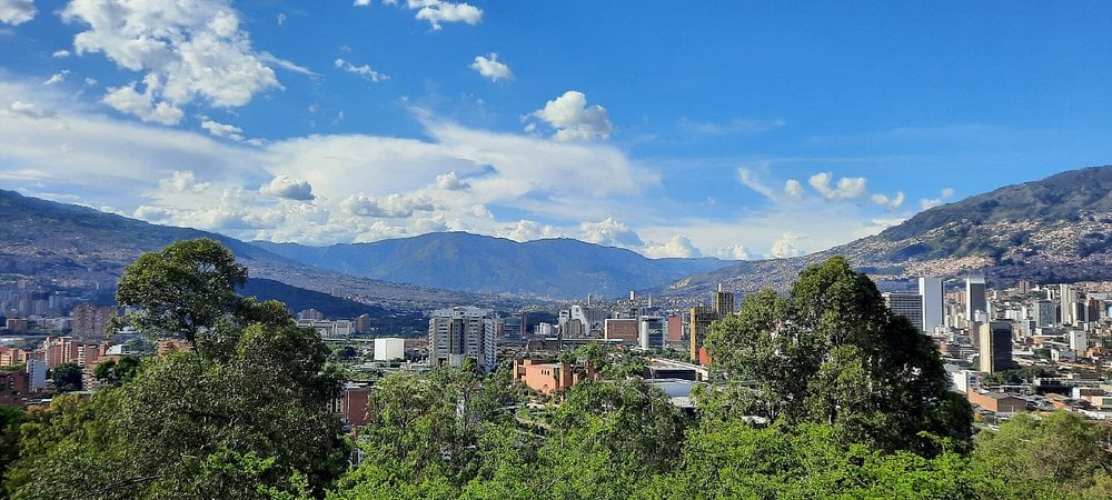 View of Medellín from Cerro Nutibara with city buildings, green trees, and surrounding mountains under a blue sky.