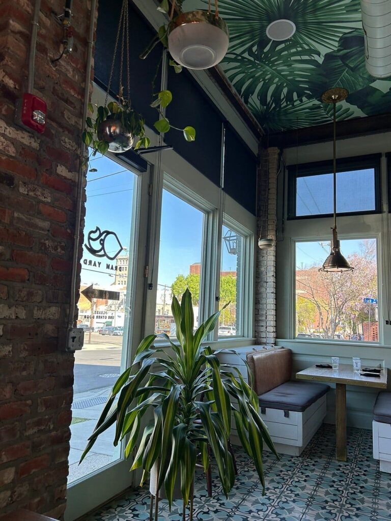 Bright corner booth at Dram Yard with large windows, leafy decor, and patterned floor tiles.