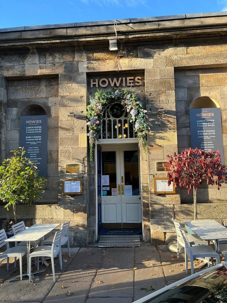 Stone-front entrance of Howies with outdoor seating and floral decor.