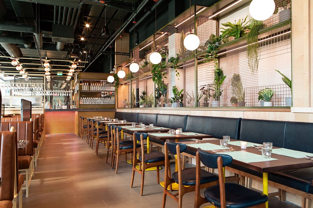 The stylish interior of Ka Pao, featuring long tables, hanging globe lights, and a wall of green plants.
