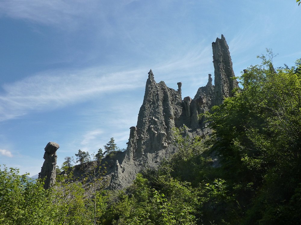 Eroded rock pinnacles of Les Demoiselles Coiffées in the French Alps, surrounded by green trees under a bright blue sky.