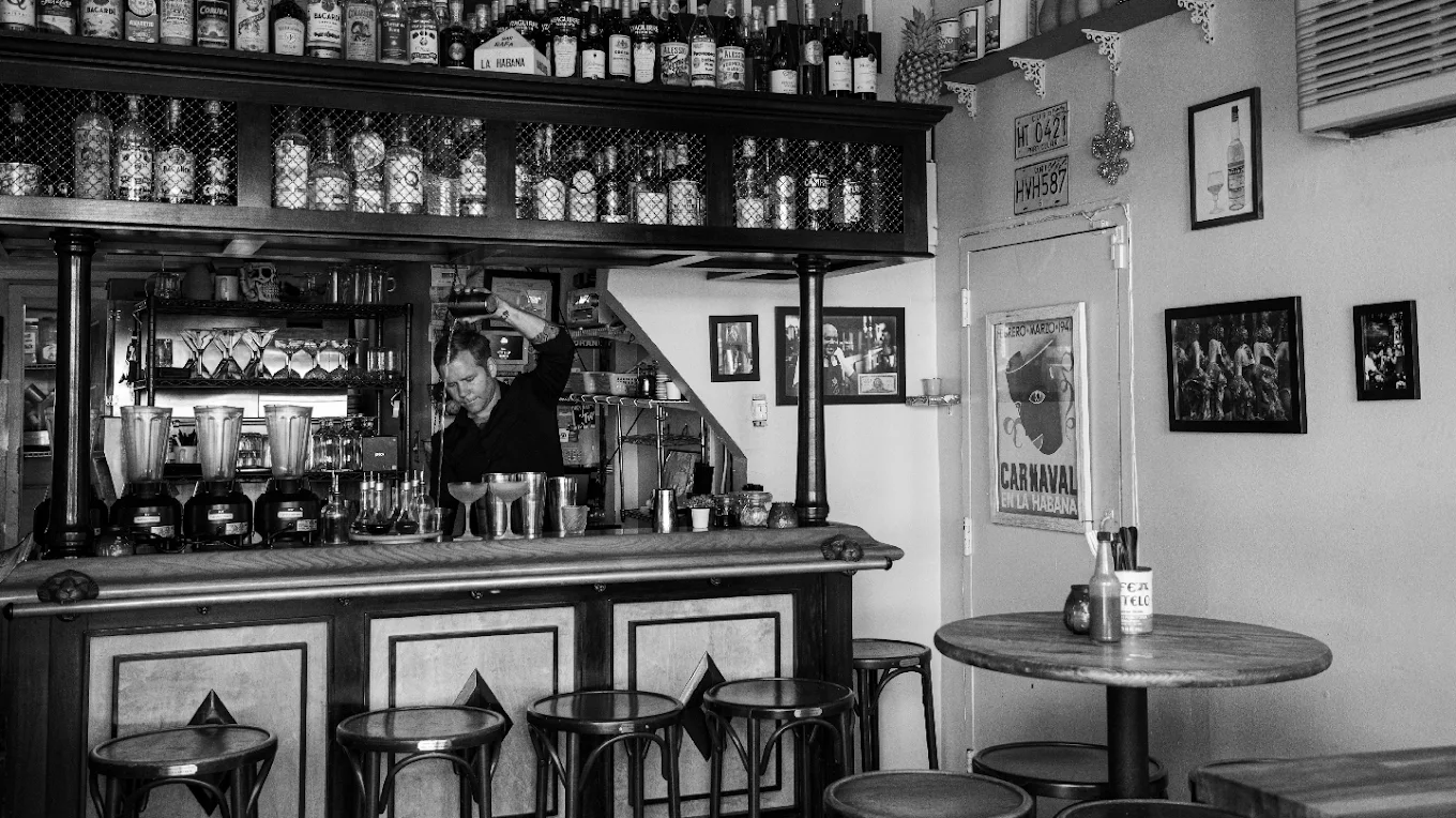 A bartender prepares cocktails behind the small bar at Manolito, surrounded by Cuban memorabilia and an extensive rum collection.