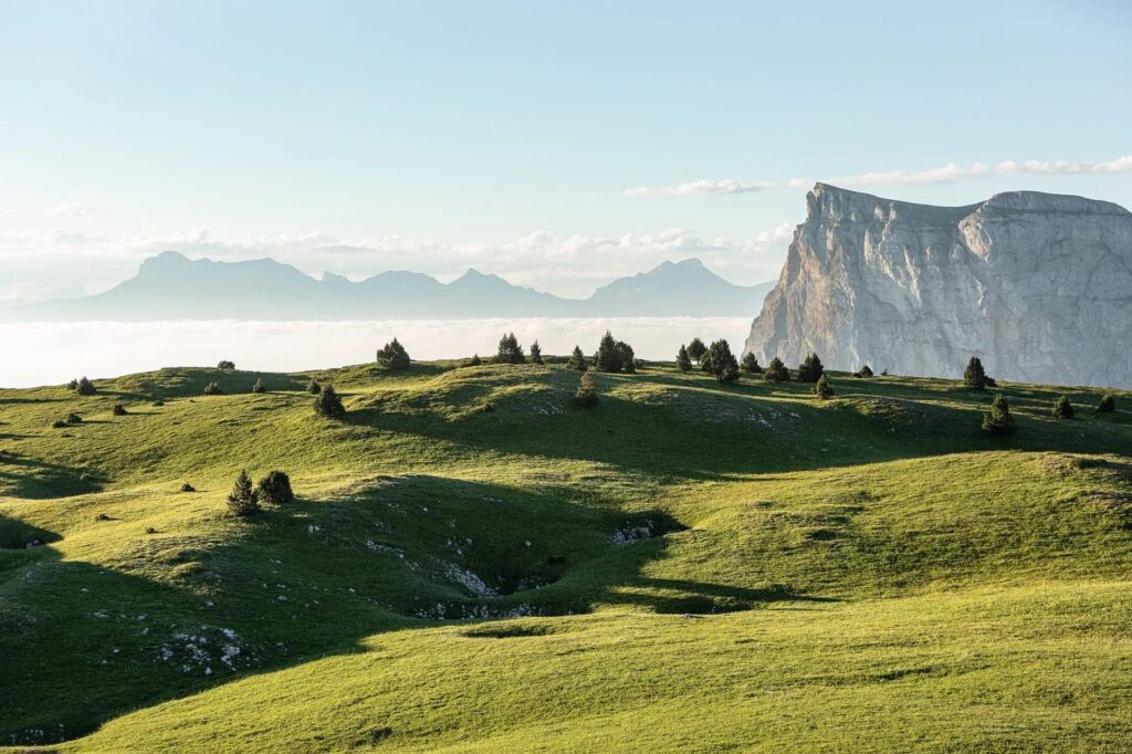 Panoramic view of Mont Aiguille in the French Alps, with rolling green hills and scattered trees under a clear blue sky.