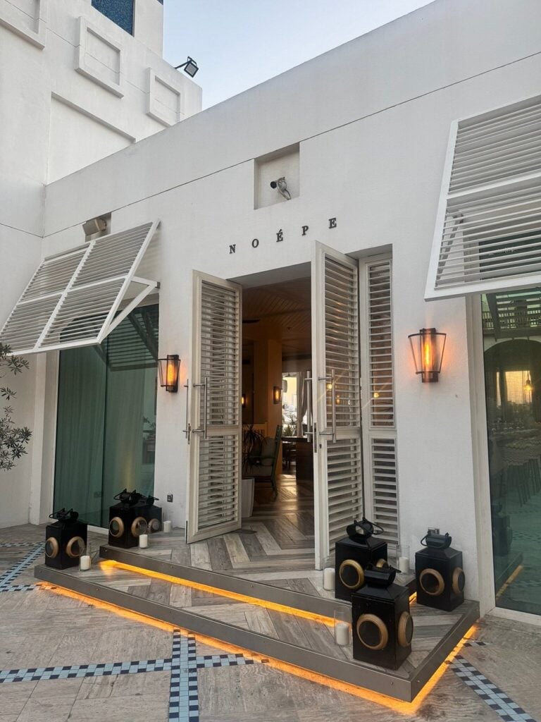 Noépe restaurant entrance with open white shutters, lanterns, and soft evening lighting.