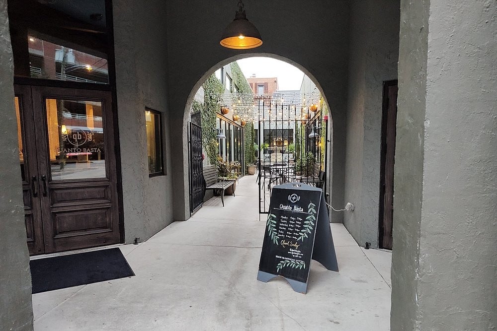 Entryway to Quanto Basta with a blackboard menu sign and outdoor seating visible through an arch.