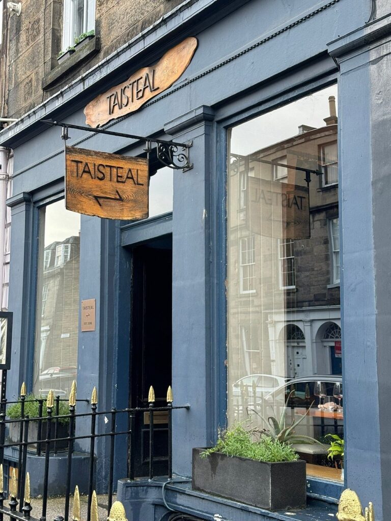 The dark blue exterior of Taisteal, with wooden signs and large windows facing the street.