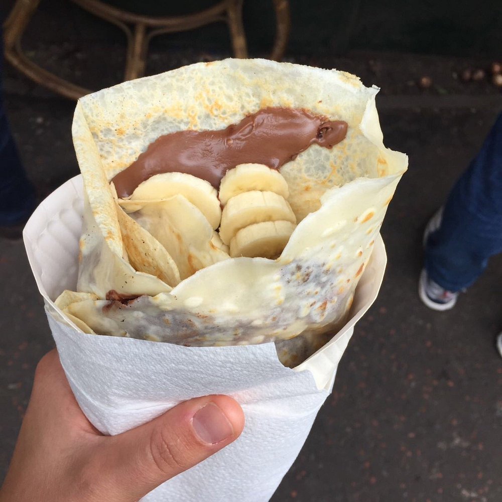 A hand holds a banana and chocolate crepe from Tupiniquim, wrapped in a paper napkin.
