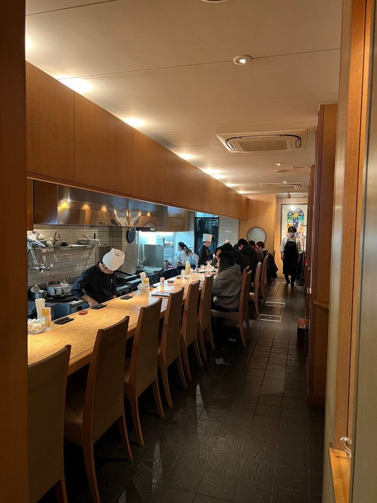 Long wooden counter at Wagyu Steak Hafuu Honten with diners seated and chefs preparing meals.