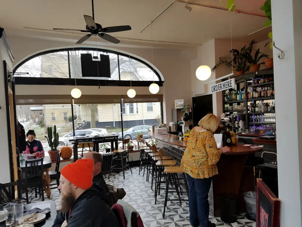 Interior view of a brightly lit cafe with a customer ordering at a wooden counter under an "Order Here" sign