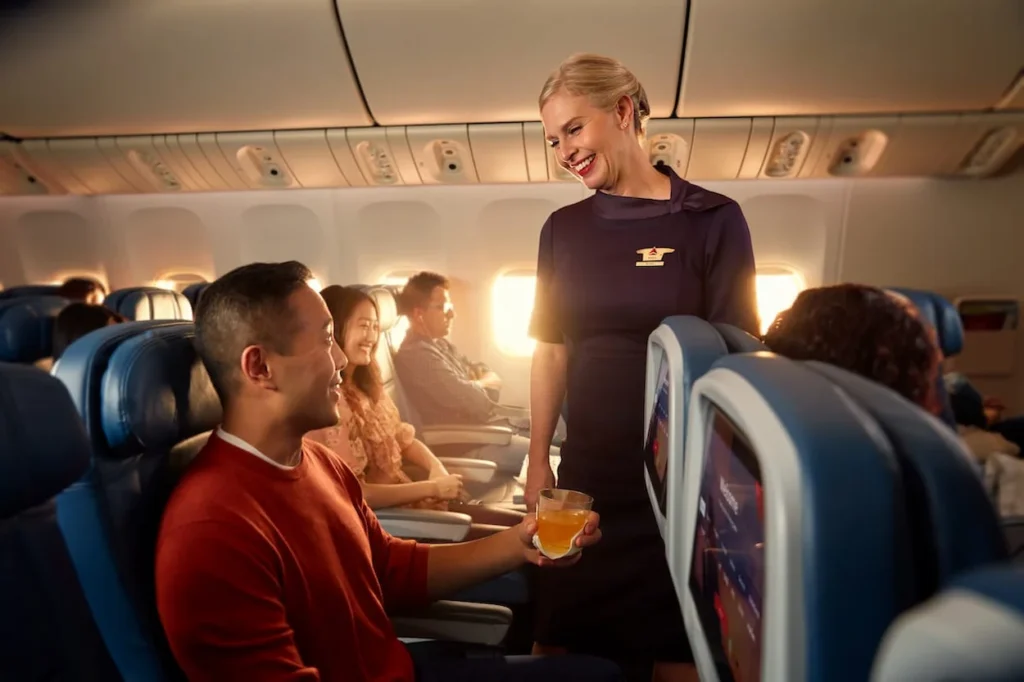 A smiling Delta flight attendant in a purple uniform serves a drink to a male passenger inside an airplane cabin