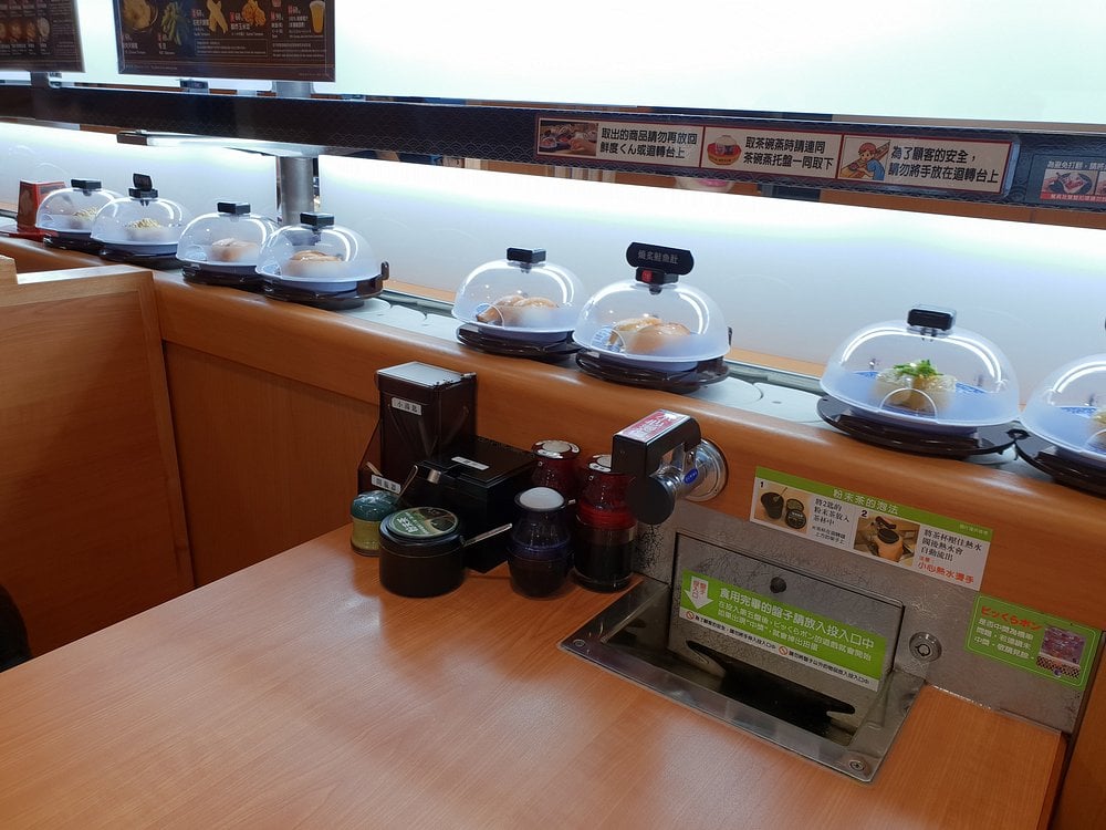 Sushi dishes on a conveyor belt at Kura Sushi with a table setup and sauce containers.
