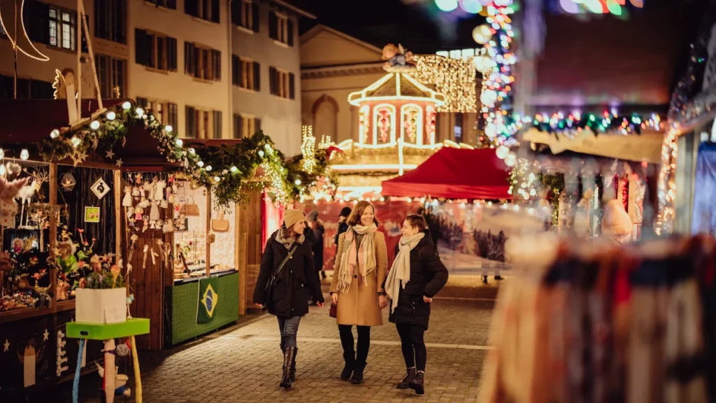 Three women walk past glowing wooden stalls at the night Winterthur Christmas Market in Switzerland