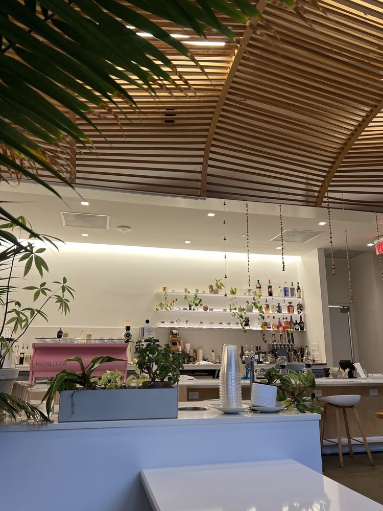 The bright, modern interior of Heirloom with a pink espresso machine, hanging wooden slats, and potted plants.