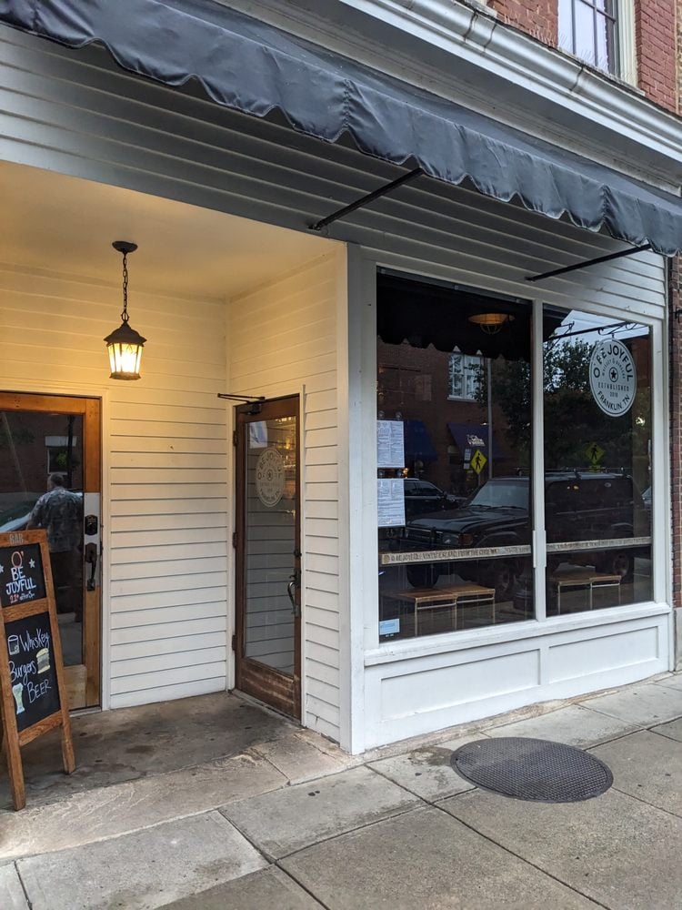Exterior of O' Be Joyful with a black awning and white siding near a sidewalk chalkboard sign.