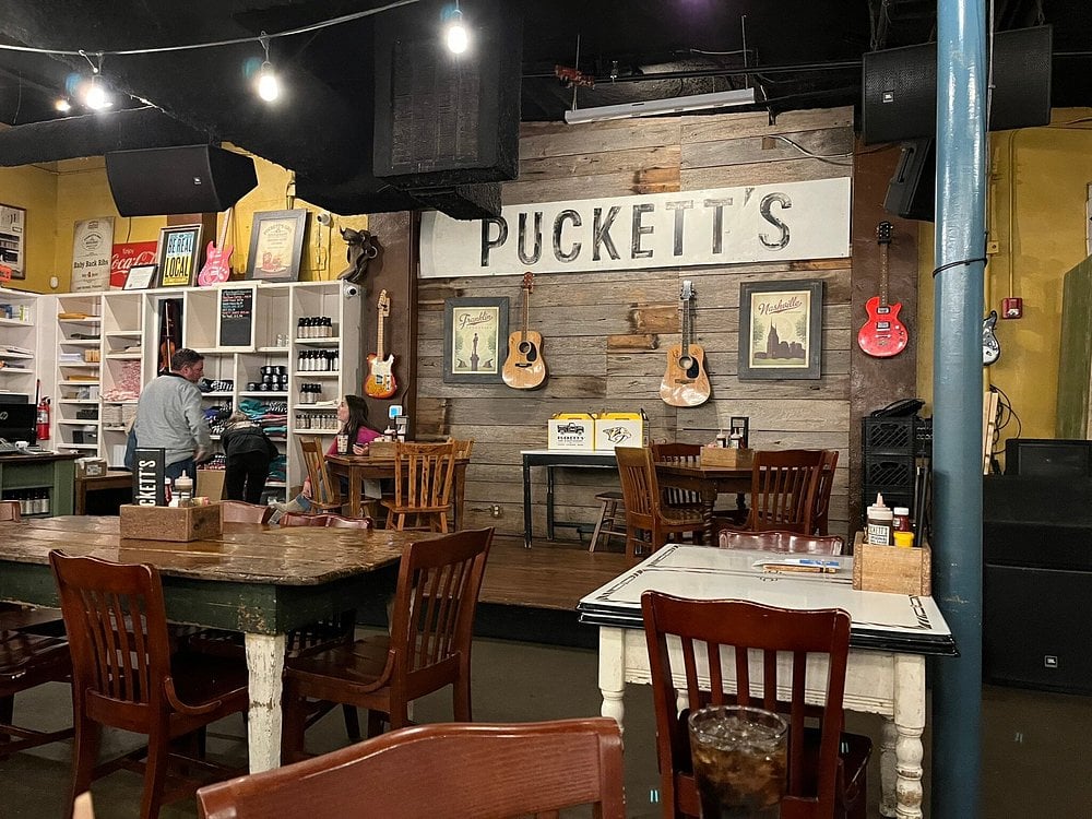 Rustic interior of Puckett's Restaurant with wooden tables and guitars hanging on a wall stage.