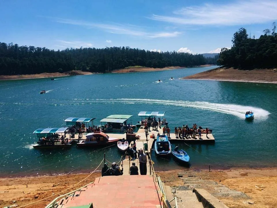 Aerial view of Pykara Lake boating dock with multiple boats, tourists, and speed boat creating wake trails on blue water surrounded by forested hills in Ooty