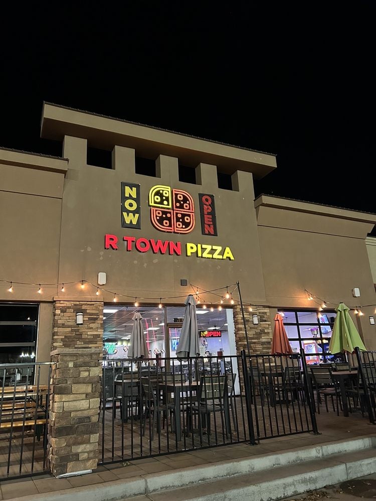Exterior of R Town Pizza at night with bright signage and patio seating under string lights.