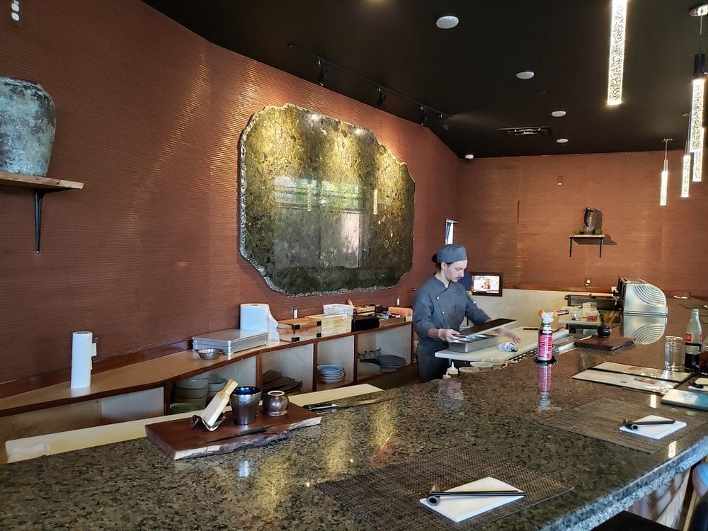 A chef prepares sushi behind the elegant stone counter at Sushi Mon, with earthy tones and ambient lighting.