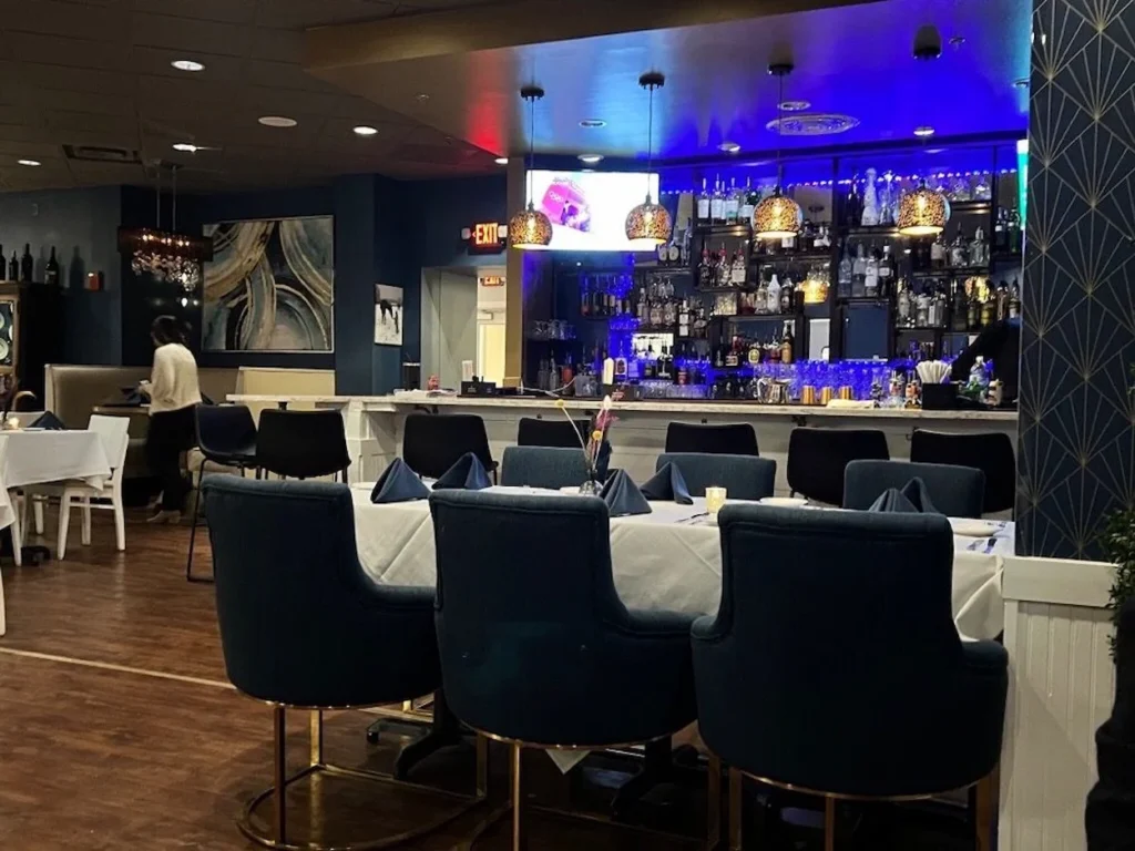 A restaurant dining room featuring a long table with blue upholstered chairs and a vibrantly lit blue bar in the background