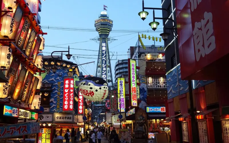 Colorful street scene in Osaka with bright signs and Tsutenkaku Tower in the background.