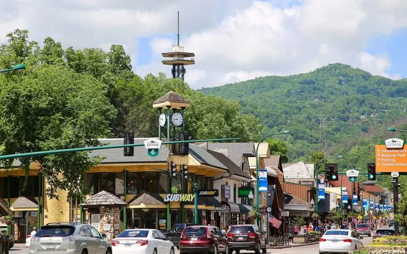 Busy Gatlinburg street with shops, traffic, and a clock tower against a mountain backdrop.