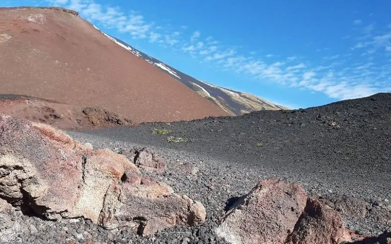 Dark volcanic rock and slopes of Mount Etna under a blue sky.