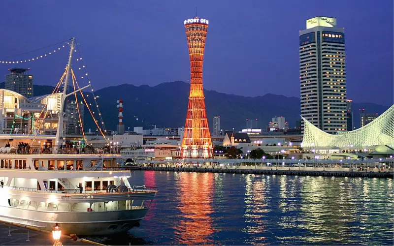 Kobe Port Tower lit up at night beside the harbor and city buildings.