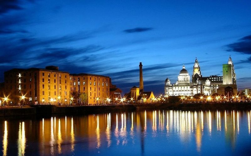 Liverpool’s waterfront at night with illuminated historic buildings reflected in the water.