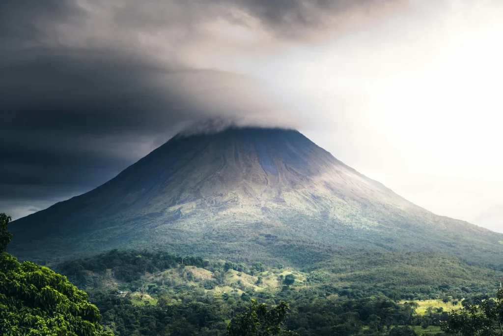 Large conical volcano peak partially covered by dark storm clouds above a green forest