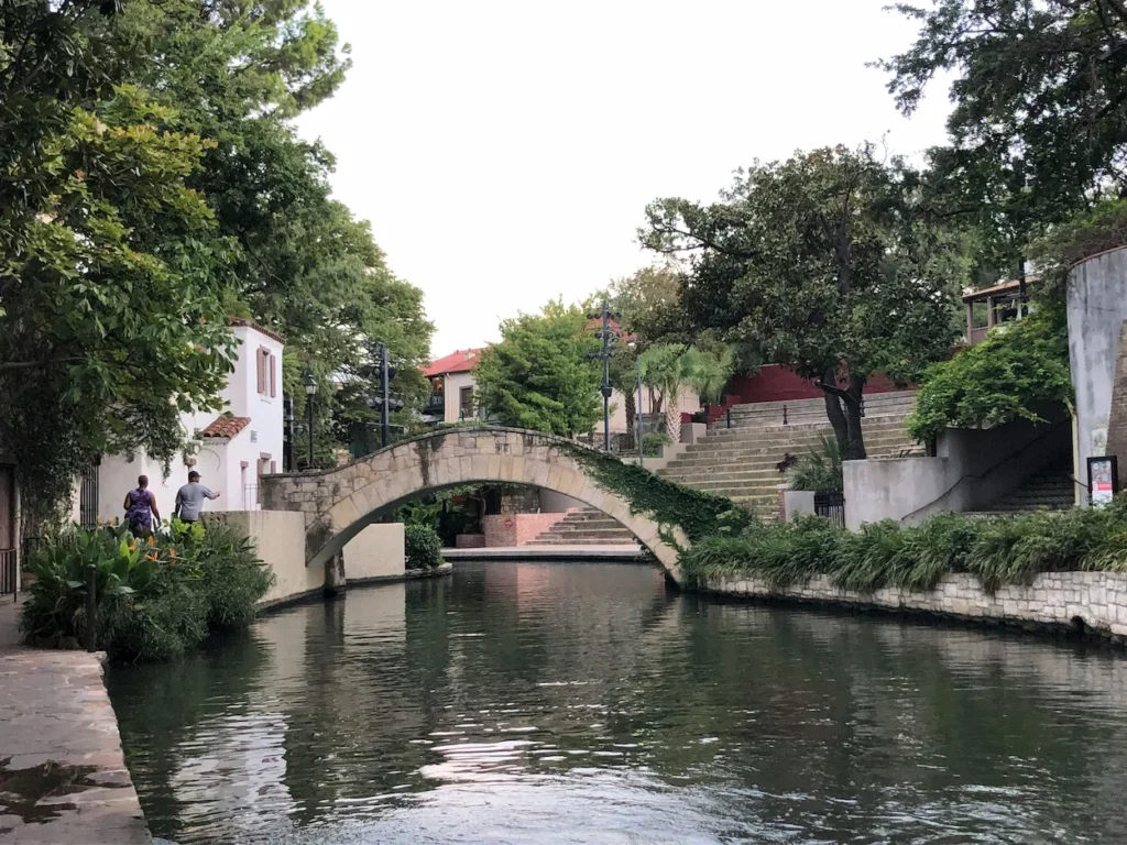 An arched stone footbridge covered in green vines stretching over the San Antonio River
