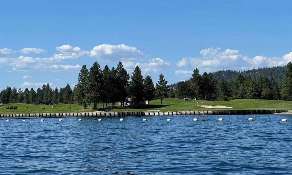 A scenic view of Coeur d'Alene Lake with trees and a golf course along the shoreline under a blue sky.