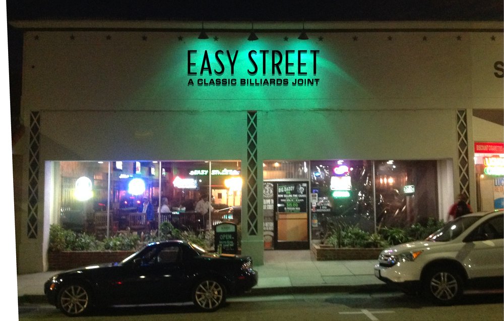 A nighttime street view of Easy Street, a classic billiards joint, with neon signs glowing and cars parked out front.