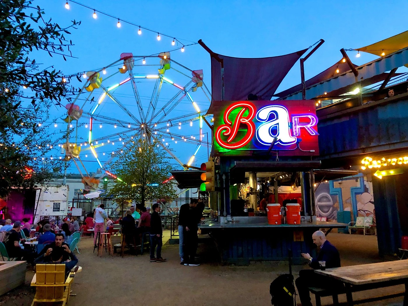 Evening scene at Truck Yard Houston showing the illuminated ferris wheel, neon bar sign, string lights, and crowds enjoying the outdoor beer garden