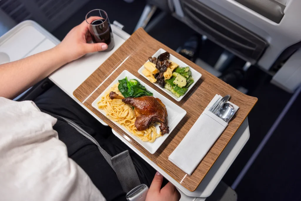 A passenger in First Class is served a gourmet duck leg and noodle meal on a wooden tray table