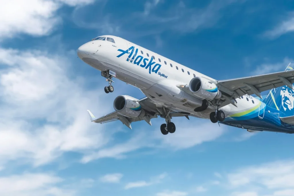 Alaska Airlines SkyWest Embraer E175 passenger airplane flying in a blue sky with white clouds during takeoff