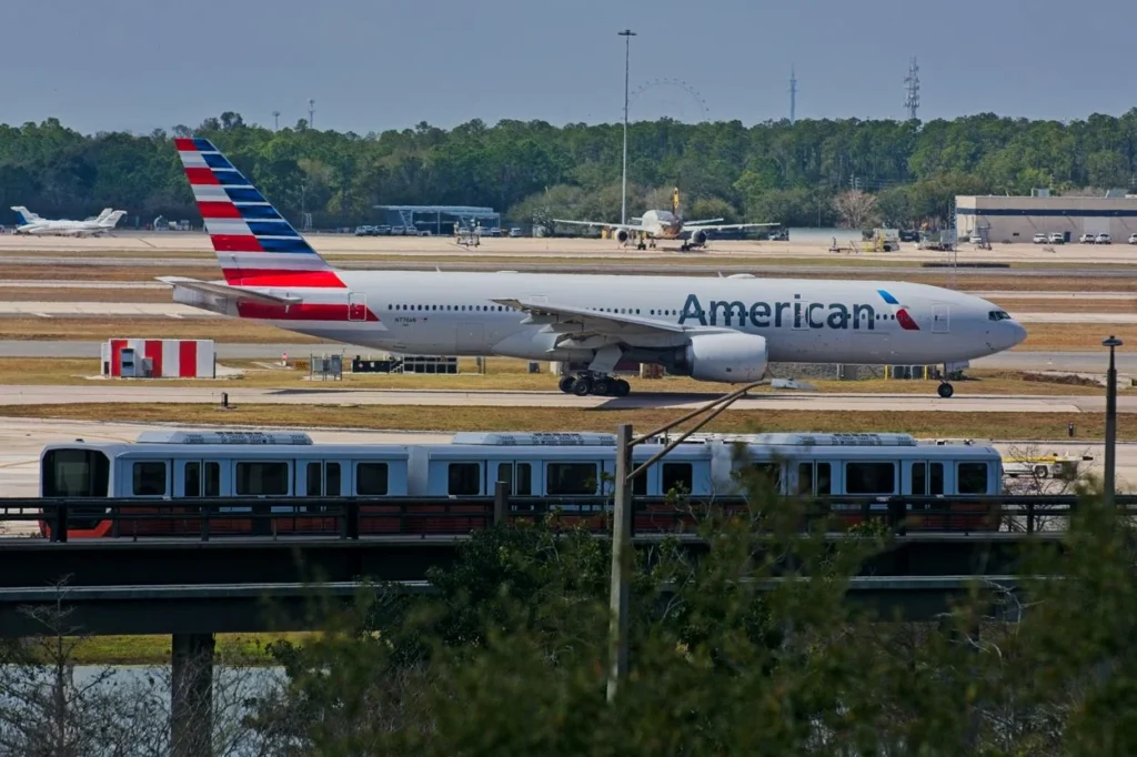 American Airlines Boeing 777 taxiing on an airport runway behind an elevated people mover train