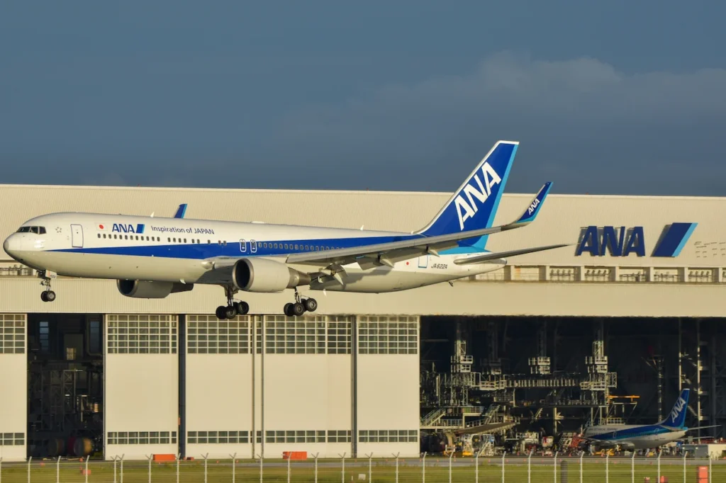ANA Boeing 767-300ER landing with landing gear down in front of a large ANA maintenance hangar