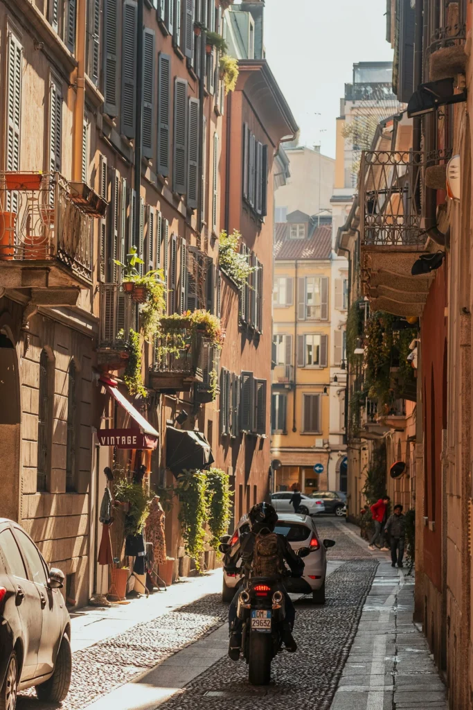 A motorcyclist riding down a narrow cobblestone street between historic apartment buildings in Milan, Italy
