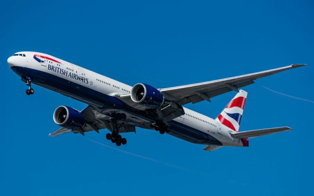 British Airways Boeing 777 passenger airliner flying through a clear blue sky with landing gear extended