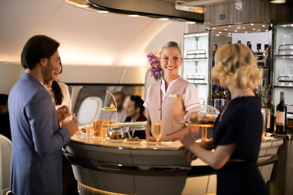 Passengers standing around a luxury onboard airplane bar with a smiling flight attendant