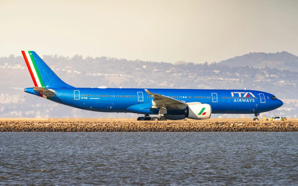 A bright blue ITA Airways Airbus A350 passenger airplane taxiing on a coastal runway next to the water