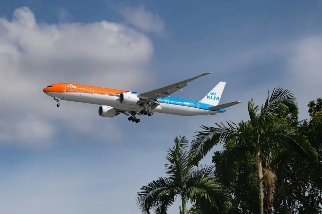 KLM Boeing 777 in orange and blue livery descending over palm trees for landing