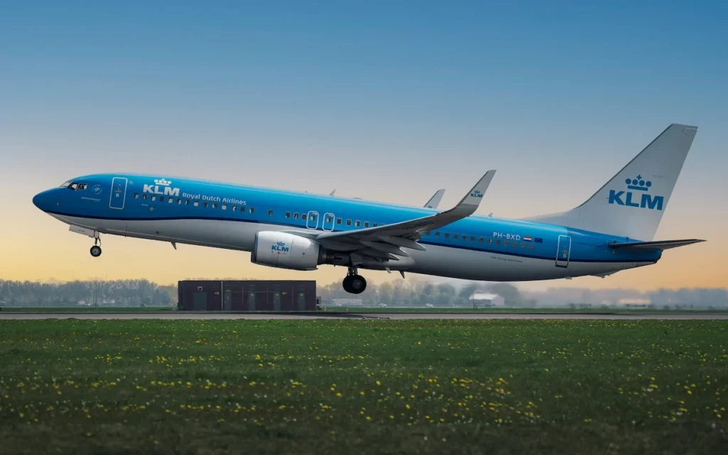 A blue and white KLM Boeing 737-800 passenger airplane taking off above a green grassy field