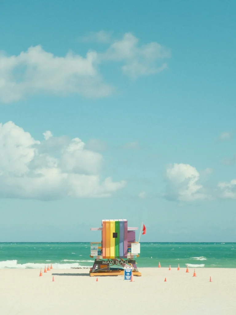 Colorful rainbow-striped Miami Beach lifeguard tower standing on white sand under a blue sky with fluffy clouds