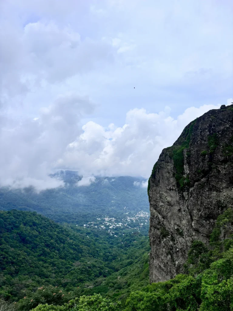 Massive steep rock cliff face overlooking a lush green mountain valley under cloudy skies
