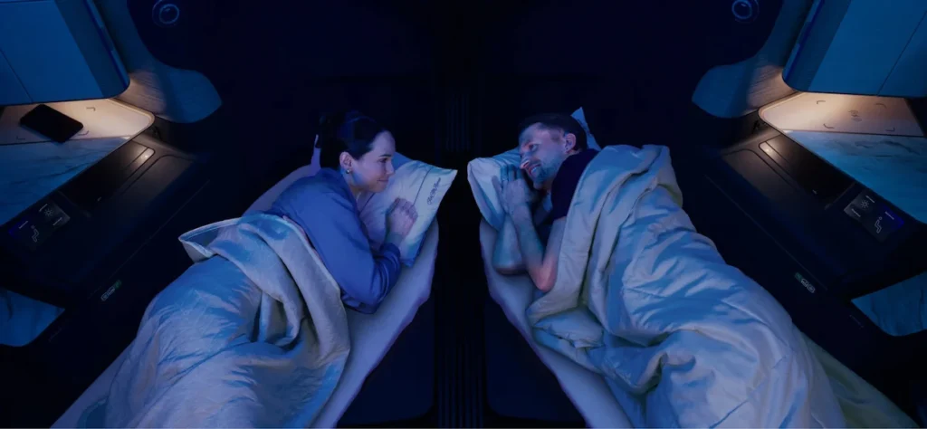Two passengers lie in adjacent airplane bed pods looking at each other under blue cabin lighting