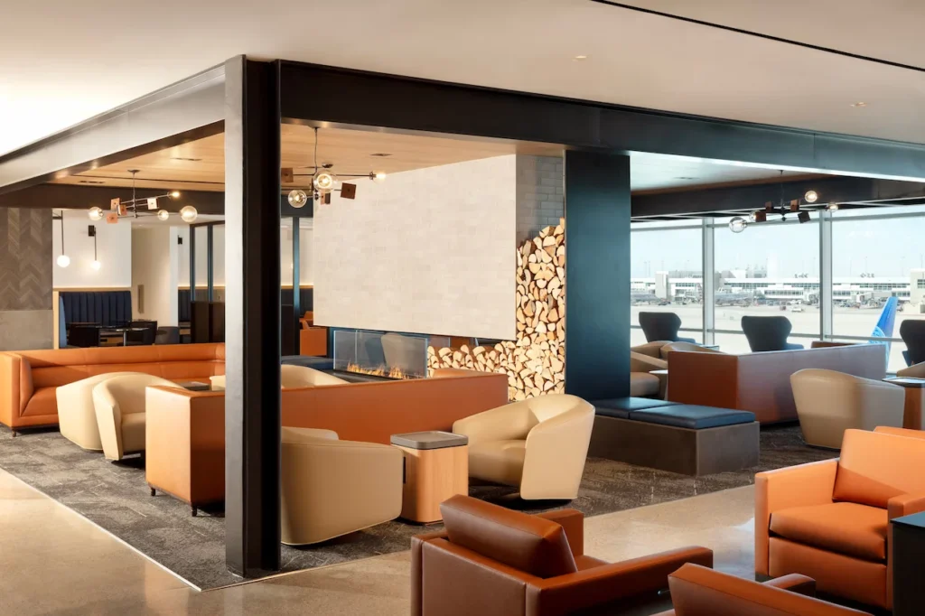 A modern airport lounge features leather armchairs, a fireplace, and views of the airfield